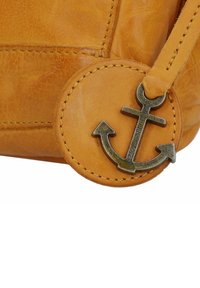 Borsa gialla in pelle con un'etichetta circolare dotata di un emblema di ancora in bronzo e dettagli di cucitura a contrasto. Texture liscia con una finitura opaca.