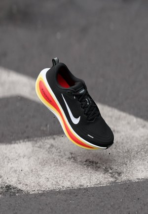 Sort løbesko med hvid swoosh, rød og gul sål, svævende over asfalt med hvide malede linjer.