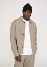 Beige Button-Down-Jacke aus einem robusten Material, mit klassischem Kragen, schwarzen Knöpfen und einem weißen T-Shirt darunter. Schwarze Mütze auf dem Kopf.