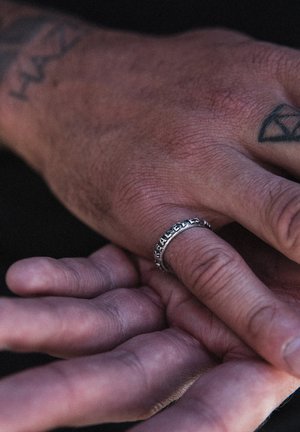 Anello in argento con design testurizzato, con lettere incise, su una mano appoggiata su un'altra mano. Toni della pelle visibili.