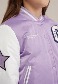 Levandų spalvos bomber style striukė su baltais rankovėmis, puošta žvaigždės aplikacija ir išsiuvinėtu užrašu. Lygus, blizgus audinys su raštuotu apykakle ir sagomis.