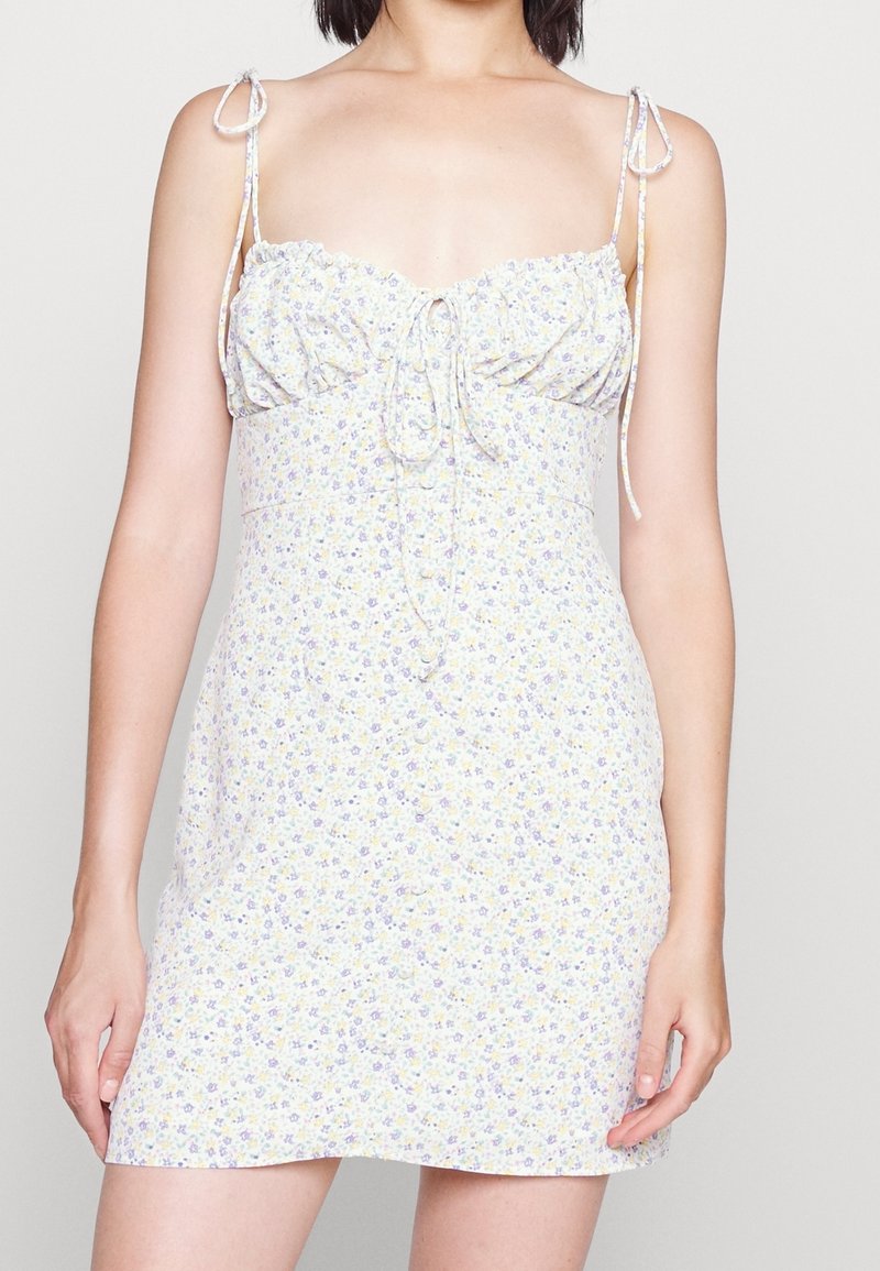 Femme portant une mini robe blanche sans manches avec un petit imprimé floral violet et jaune et des bretelles nouées sur les épaules.