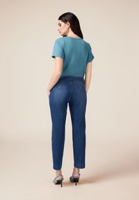 Camisa de manga corta color verde azulado combinada con jeans ajustados de color azul oscuro que tienen bolsillos y una textura suave, llevada con sandalias de tacón grises.