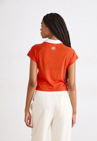 Rode katoenen top met korte mouwen, een witte kraag en een logo op de rug. In combinatie met lichtbeige gestreepte broek.