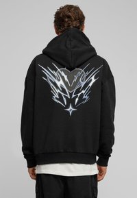 Mustat hupparit, joissa on suuri metallinhohtoinen hopeinen graafinen kuva, joka esittää sydäntä ja liekin kaltaisia elementtejä selässä. Puuvillasekoitetta, rento malli.