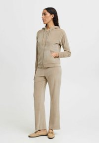 Beige sametový sportovní komplet s mikinou na zip, která má přední kapsu, a odpovídající kalhoty s uvolněným střihem. Měkká textura, minimalistický design.