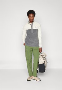 Fleece pullover in het wit en grijs, met een kraag en knopen. Groene cargo broek. Beige en zwarte rugzak met een gekreukt bovenstuk, persoon houdt deze vast.