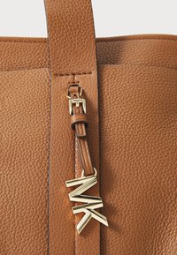 Bruine leren tote met structuur en een verwijderbare bruine schouderband, versierd met een gouden MK-logo charm. Kenmerkt zich door strakke lijnen en een gestructureerd ontwerp.