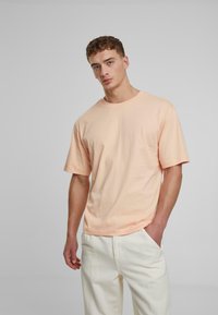 Kurzärmliges T-Shirt in Pfirsichfarbe aus Baumwolle. Entspannte Passform mit Rundhalsausschnitt und nahtlosen Schultern. Kombiniert mit hellen Hosen.