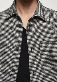 Chaqueta de patrón de espiga en blanco y negro con cuello con solapa, cierre de botones y un bolsillo delantero. El material parece texturizado.
