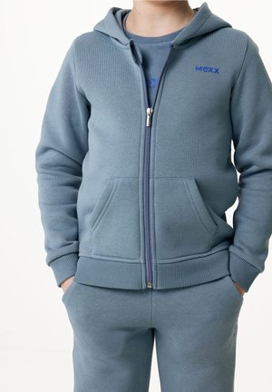 Kind trägt blauen Kapuzenpullover mit Reißverschluss und passende Jogginghose, Hände in den Taschen, Gesicht nicht sichtbar.