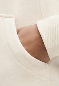 Sweatshirt de culoare crem cu un buzunar frontal, manșete ribbate și textură netedă. Materialul pare moale, cu detalii de cusătură subtile.