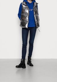 Gilet in piuma argentato con finitura lucida, indossato sopra un felpa blu. Abbinato a jeans scuri e stivaletti militari neri, con zip e fibbie.