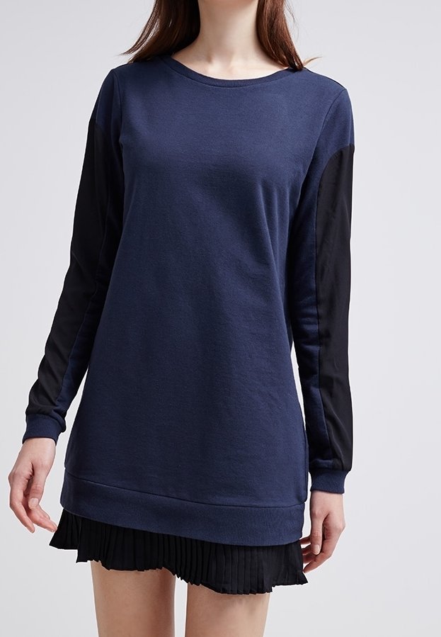 Woman wearing a long navy blue sweatshirt with black contrasting sleeves over a black pleated skirt, standing against a plain background.