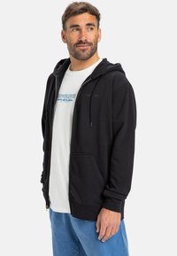 Sweatshirt preto com fecho éclair, dois bolsos frontais, capuz com cordão e logótipo bordado. Usado por cima de uma t-shirt branca e combinado com calças azuis.