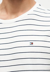 Κοντινό πλάνο ενός ατόμου που φοράει λευκό T-shirt με λεπτές οριζόντιες ναυτικά μπλε ρίγες και μικρό ορθογώνιο κόκκινο και ναυτικό μπλε λογότυπο στο στήθος.