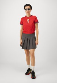 Red ribbed Nike jersey with blue accents and emblem, paired with a grey pleated skirt and black footwear featuring neon details.