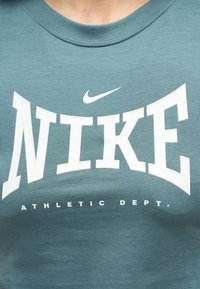 Κοντινή λήψη μπλουζάκι σε τιρκουάζ χρώμα με μεγάλο λευκό κείμενο "NIKE", λευκό σύμβολο Nike από πάνω και την επιγραφή "ATHLETIC DEPT." από κάτω.