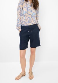 Shorts en lin bleu marine avec taille à cordon, deux poches latérales, accompagnés d'un chemisier léger à fleurs avec des contours bleus sur beige.