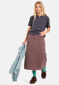Grå t-shirt, brun lang nederdel, grønne sokker, brune sko, holder en blå og grønternet skjorte. Enkel, afslappet påklædning med lagdelte teksturer.