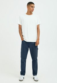White crew-neck t-shirt paired with navy cargo pants, featuring pockets. Model wears white and gray sneakers. Simple, casual outfit.