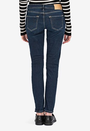 Personne portant un jean skinny bleu foncé avec des poches arrière, un haut à manches longues noir et blanc à rayures, et des mocassins noirs, debout dos à nous.