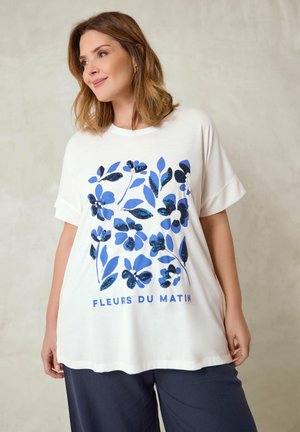 Woman wearing a white oversized t-shirt with blue floral design and "Fleurs du Matin" text, paired with navy blue pants against a light background.