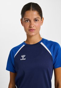 Tumejate juustega naine, kellel on selja taha tõmmatud juuksed, kandes tumesinist ja kuninglikku sinist spordisärki valge Hummeli logoga rinnal.