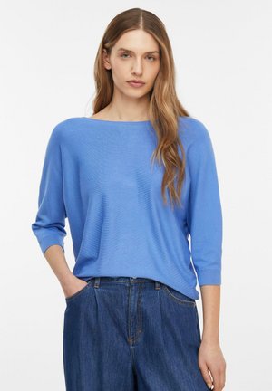 Kobieta o długich jasnobrązowych włosach, ubrana w jasnoniebieską bluzkę z długim rękawem i ciemnoniebieskie, wysokie dżinsy z jedną ręką w kieszeni.