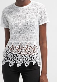 Top bianco in pizzo con maniche corte, caratterizzato da un delicato motivo in crochet floreale all'orlo, sovrapposto a una parte inferiore nera uniforme.