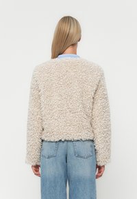 Pullover corto color beige chiaro e fluffy con un tessuto texture. Presenta maniche lunghe e un collo rotondo, abbinato a jeans azzurri.