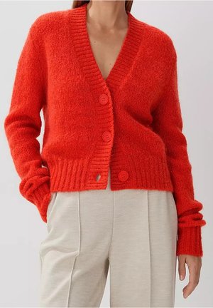 Woman wearing a bright red buttoned cardigan with long sleeves and light beige pants, standing with one hand in pocket.