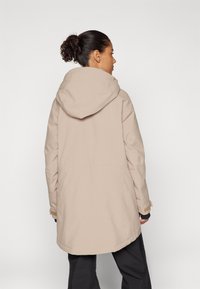 Chaqueta impermeable beige con capucha, que presenta un corte holgado y costuras visibles. Los puños tienen acentos de tela ajustables.