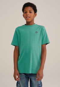 Žalias medvilnės marškinėliai trumpomis rankovėmis, apvaliu kaklu ir maža dekoratyvine fixacija ant kairės krūtinės. Lygi tekstūra su laisvu kirpimu.