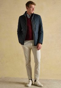 Námornícky prešívaný kabát s vysokým golierom, burgundský sveter, svetlosivé chinos nohavice a biele tenisky. Hladká textúra a ležérny dizajn.