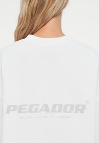 Valge dressipluus, millel on ümar kaelus ja suur heledat halli värvi "PEGADOR" logo trükitud seljale. Pehme kangas, millel on sile tekstuur.