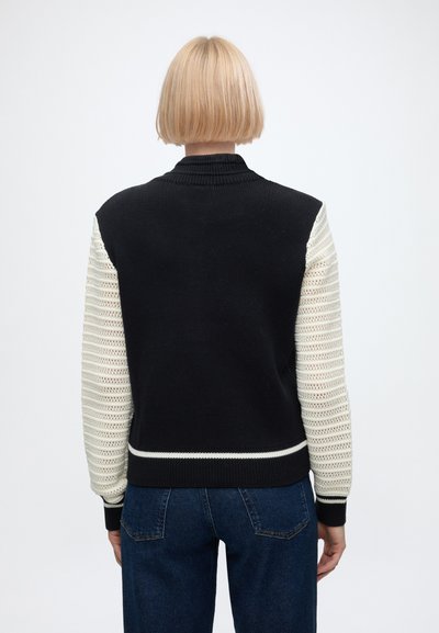 Personne aux cheveux blonds courts porte un gilet noir en maille avec des manches longues blanches tricotées et un jean bleu, vue de dos sur un fond uni.