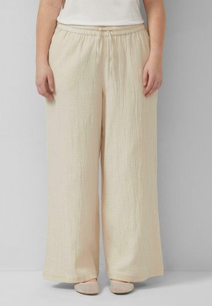 Femme portant un pantalon large crème texturé à taille haute et taille élastique, accompagné de chaussures plates assorties, debout devant un fond uni.