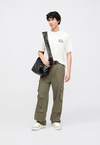 Hvid T-shirt med grafik, olivengrønne cargobukser med lommer, sort skuldertaske og lyse sneakers. Modellen står foran en ensfarvet baggrund.