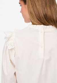 Blusa blanca con escote con volantes, detalles de encaje en los hombros, cierre de botón en la parte posterior y una textura de tejido suave y ligera.