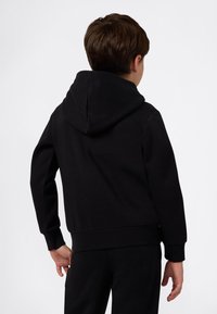 Jongen staat met zijn rug naar de camera gekeerd, draagt een zwarte hoodie en bijpassende zwarte broek op een effen achtergrond.