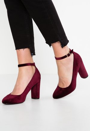 Pieds chaussés de talons blocs en velours rouge foncé avec bride à la cheville, assortis à un jean skinny noir coupé court avec des ourlets effilochés.
