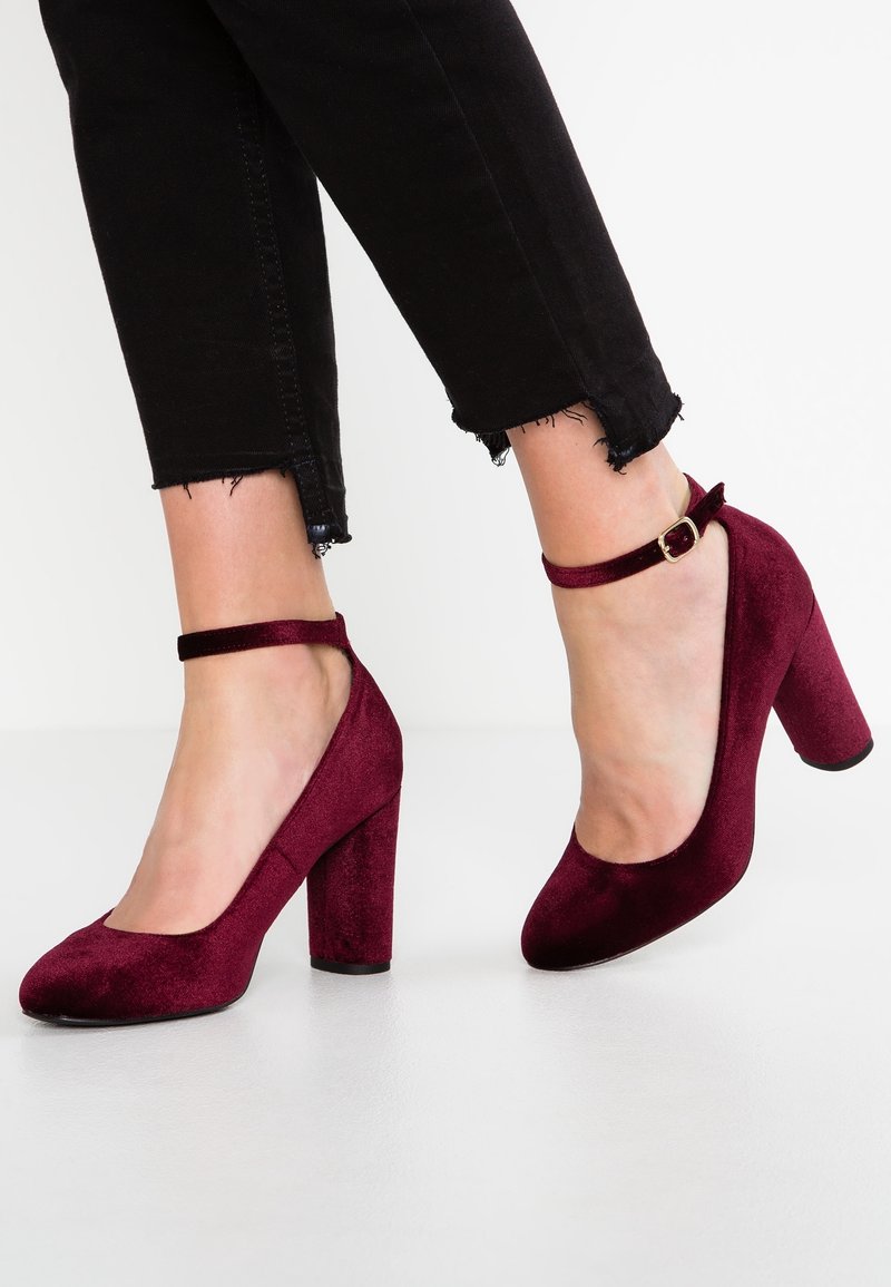 Pieds chaussés de talons blocs en velours rouge foncé avec bride à la cheville, assortis à un jean skinny noir coupé court avec des ourlets effilochés.