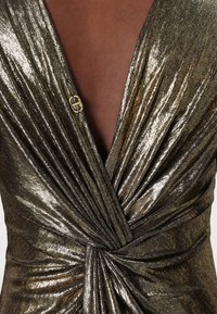 Metalická zlatá a bronzová zavinovací šaty s vertikálními sklady, hlubokým výstřihem do V a dekorativní zlatou logovací sponou na hrudi.