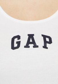 White ribbed tank top featuring a black "GAP" logo on the front. The neckline is scooped, and the fabric has a soft texture.