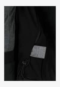 Interior textil gris con un forro negro, que presenta una etiqueta de cuidado y un ajuste con cordón. Textura suave con un detalle de cremallera.