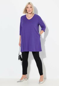 Lila Tunika-Oberteil mit Dreiviertelärmeln, kombiniert mit schwarzen Leggings. Akzentuiert mit silbernen High Heels und einer kleinen schwarzen Handtasche.