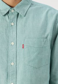 Camicia button-up in tessuto color turchese chiaro, con taschino sul lato sinistro e un'etichetta rossa. Texture liscia e design del colletto standard.