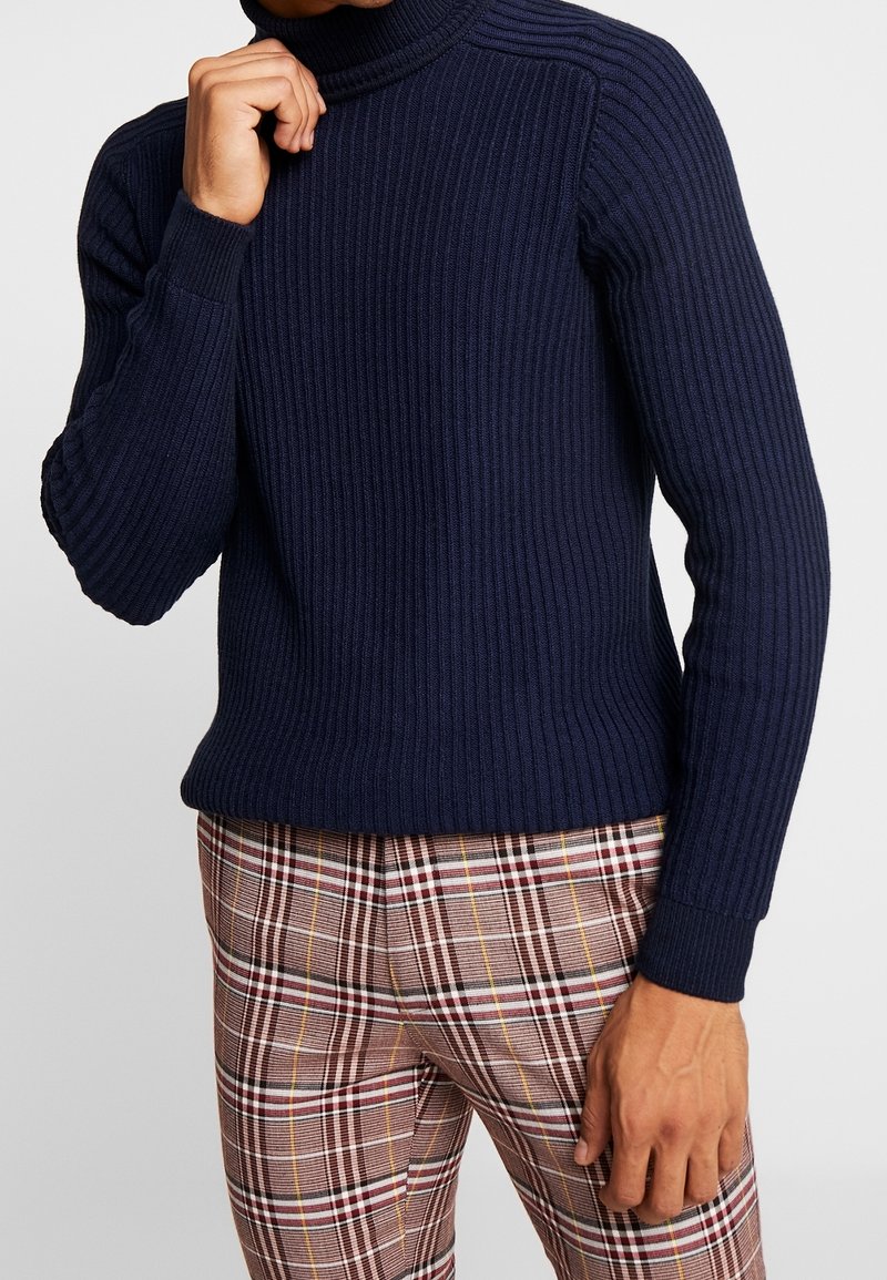 Homme portant un pull à col roulé côtelé bleu marine et un pantalon à carreaux marron, une main près du menton et l'autre détendue.