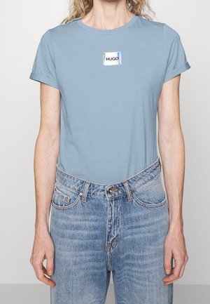 Hellblaues Baumwoll-T-Shirt mit kurzen Ärmeln und einem rechteckigen Etikett mit dem "HUGO"-Logo. Kombiniert mit hoch taillierter, ausgewaschener blauer Jeans.